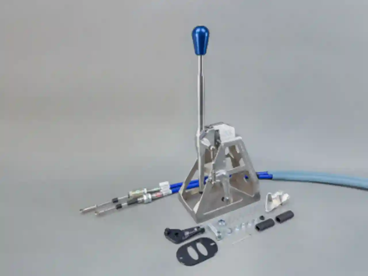 Studio shot of a CAE Ultra Shifter made of silver-colored aluminum with a blue shift knob, accessories, and shift cables for the Ford Focus MK1, set against a gray background.