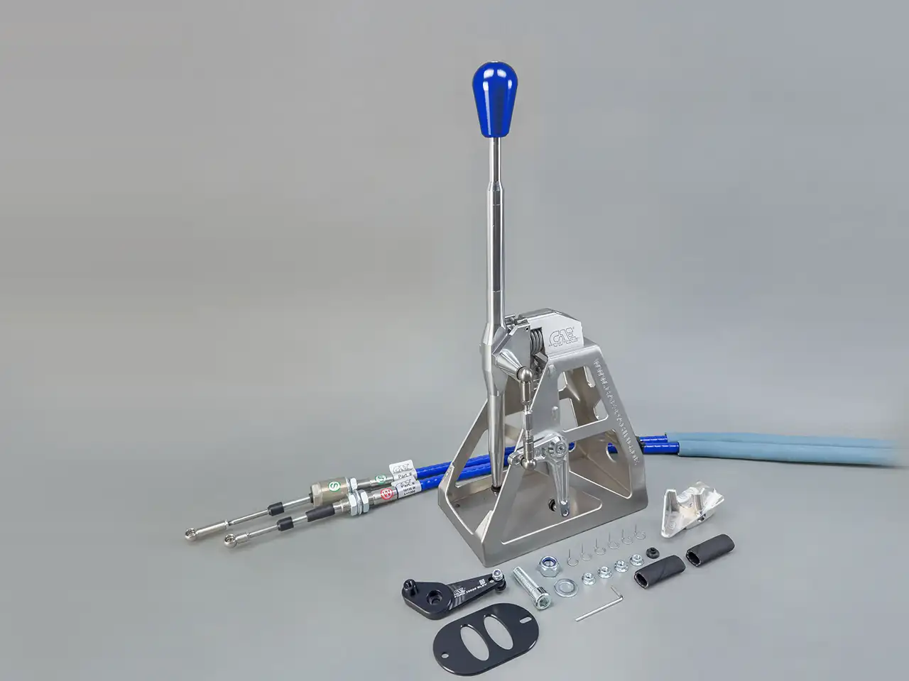 Studioaufnahme eines CAE Ultra Shifters aus silberfarbenem Aluminium mit blauem Schaltknauf, Zubehör und Schaltseilen für dem Ford Focus MK1 auf grauem Hintergrund.